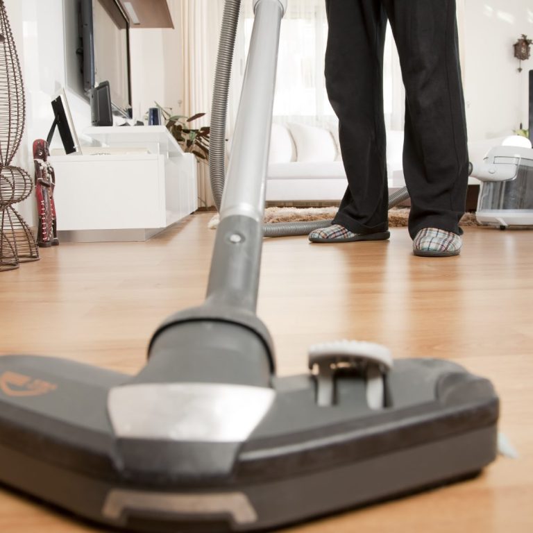 Un aspirateur est utilisé sur un sol en bois, avec des pieds en pantoufles visibles.